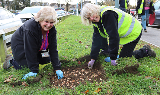 Two volunteers wearing high vis jackets planting bulbs at the front of the RUH and smiling at the camera