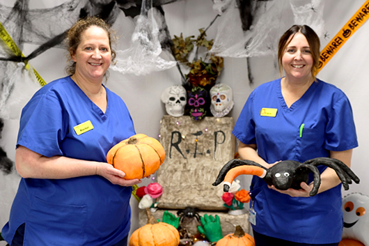 Claire and Rachel from the orthopaedic team standing in front of the halloween display which features spider webs, a tombstone, pumpkins and a spider.  They are both smiling.